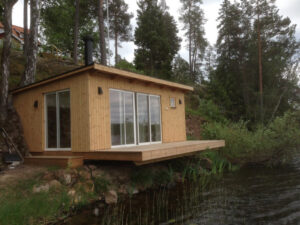 Bastuhus i trä med stora glaspartier byggt vid en sjö, omgivet av skog.